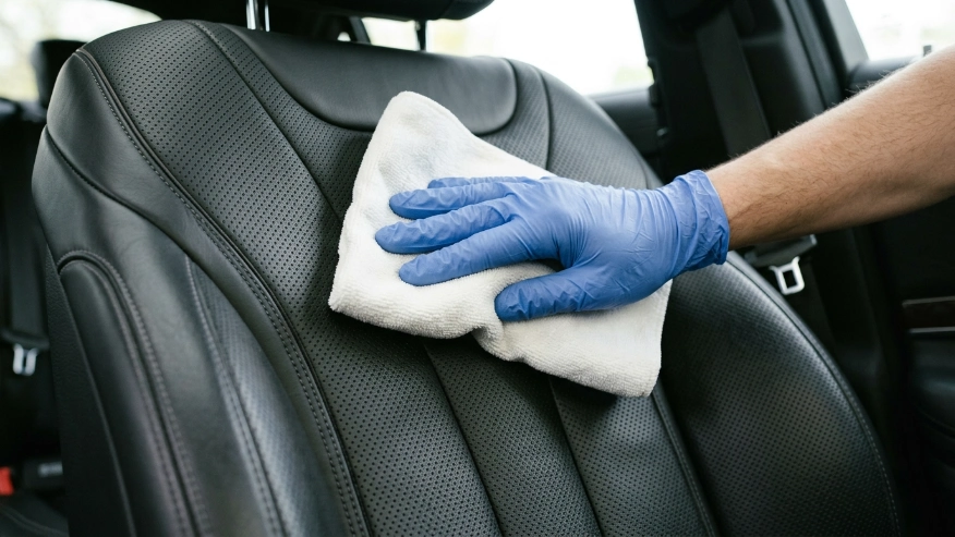 A person's hand using a white microfiber cloth to gently wipe a premium leather car seat.